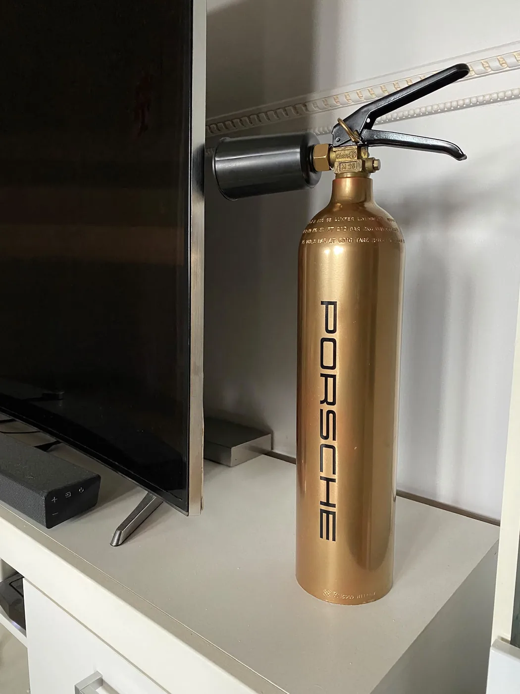 Extincteur CO2 doré personnalisé au lettrage noir Porsche, posé sur un meuble TV blanc — Art'cade Design Nice