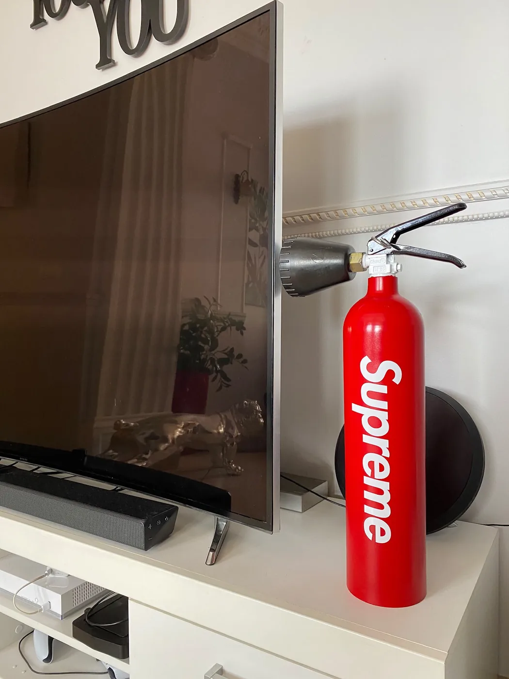 Extincteur rouge personnalisé au logo blanc Supreme, posé sur un meuble TV blanc — Art'cade Design Nice