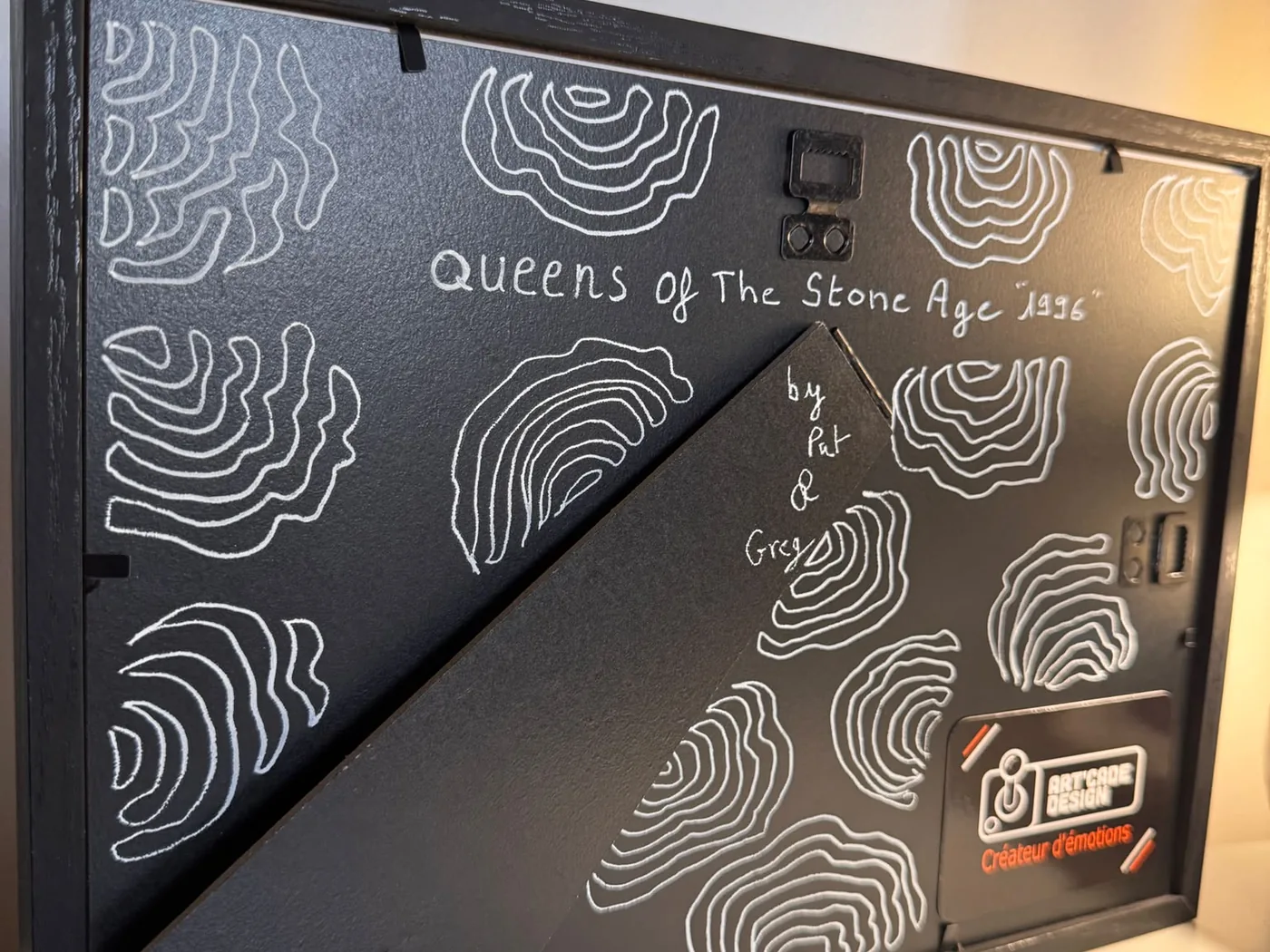 Le dos d'un cadre terminé : motif de lignes blanches tracées à la main sur carton noir, la mention manuscrite « Queens of The Stone Age "1996" » et « by Pat & Greg », et l'étiquette Art'cade Design « Créateur d'émotions ».
